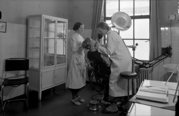 02 Foto: Teigens Fotoatelier / DEXTRA Photo, Teknisk museum. Bildetekst: Et barn sitter i tannlegestolen hos skoletannlegen ved Hersleb skole. Kvinnelig tannlege og tannlegeassistent