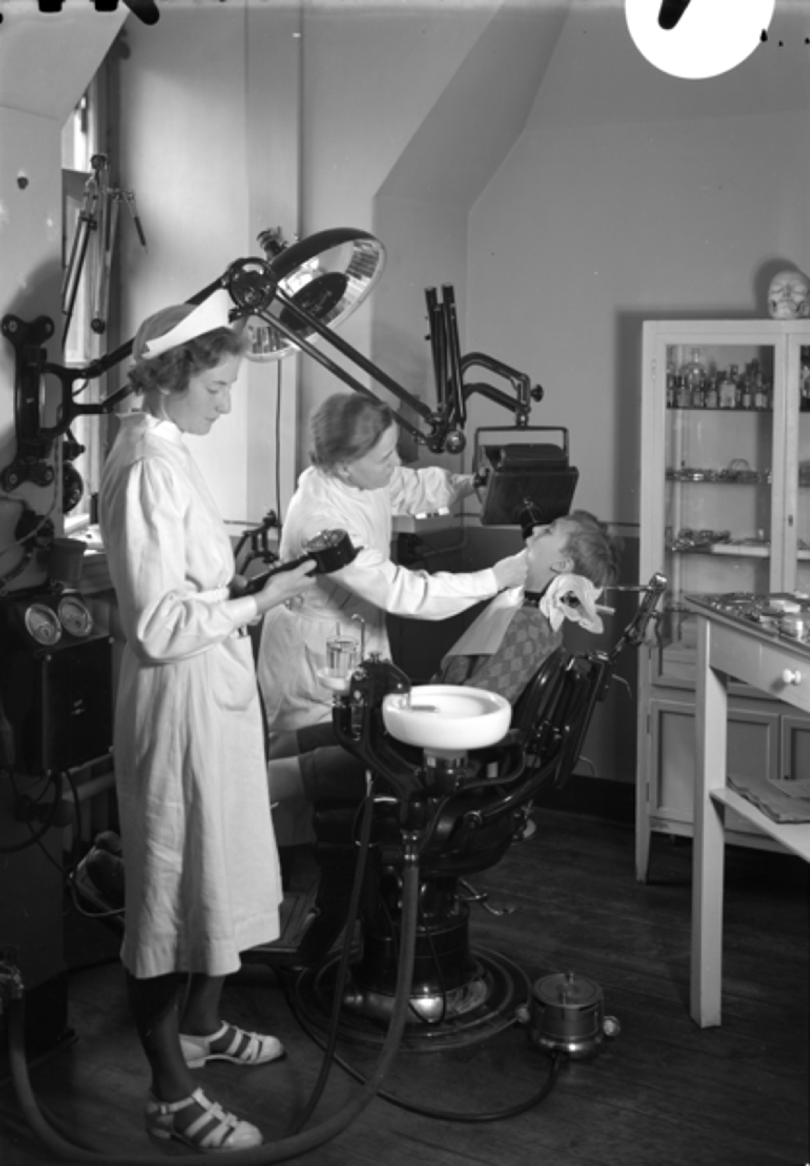 03 Foto: Teigens Fotoatelier/ DEXTRA Photo, Teknisk museum. Bildetekst Et barn sitter i tannlegestolen hos skoletannlegen ved Hersleb skole. Kvinnelig tannlege og tannlegeassistent. .I fotografens arkiv står det: Skoletannlege, Hersleb skole, Dr. Gytfe