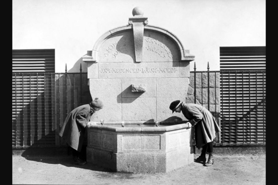 14 Fotograf ukjent, 1922. Byarkitekten. Bildenummer: A-20031/Ua/0003/007.Utlånt av Oslo byarkiv. Bildetekst: Drikkefontene, to jenter som drikker vann. "I MIMERS BRØND ER VANDET KOLDT. FRA HEDENHOLD I ÆRE HOLDT". Herslebs skole 1922